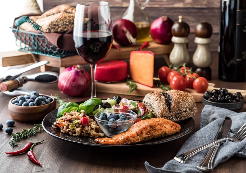 Mediterranean meal of greek salad, seven grain salad, salmon fillet and a small bowl of blueberries, surrounded by Mediterranean diet foods.